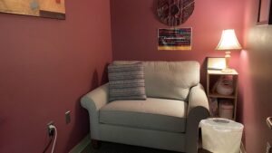 The interior of the Maxine Tracy Wellness Room hosts a comfortable gray couch, muted red walls, soft warm lighting, a trashbin, and shelf with a sound machine and breast pump.