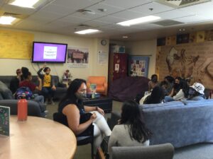 Community members seated around the couches in the Women's Center lounge participating in an orientation activity! A slideshow on the TV reads "First, pair up with someone you do not already know. Now, find one commonality between you two..."